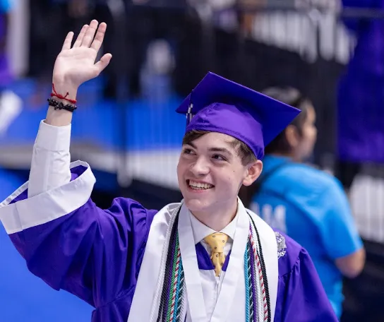 Austin ISD graduate in purple cap and gown, smiling and waving